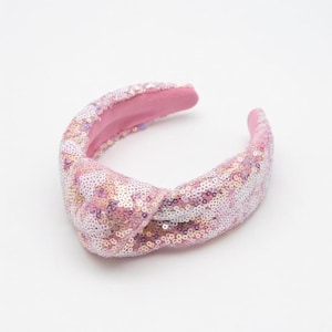 Pode incluir: Uma tiara rosa com lantejoulas e design de nó. A tiara apresenta uma mistura de lantejoulas rosa, brancas e iridescentes. O interior é forrado com um tecido rosa liso, um acessório de cabelo elegante.