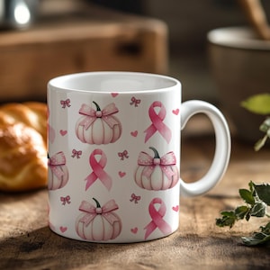 May include: White ceramic mug featuring a repeating pattern of pink pumpkins with bows, pink ribbons, and small pink hearts. The mug has a white handle and is set on a wooden surface.
