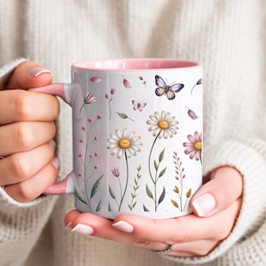 Puede incluir: Taza de cerámica blanca con interior y asa rosas, adornada con un diseño floral con margaritas, mariposas y otras flores silvestres. La taza está sostenida por una persona que lleva un suéter blanco.