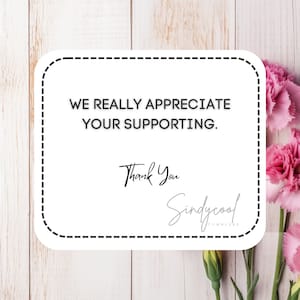 Peut inclure: Une carte blanche avec une bordure noire en pointill&eacute;s et le texte "We really appreciate your supporting. Thank You Sindycool Tumblers". La carte est sur un fond en bois blanc avec des fleurs roses.