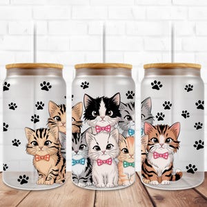 Könnte beinhalten: Drei klare Glasbecher mit Bambusdeckeln und Strohhalmen. Jeder Becher zeigt ein anderes Design mit niedlichen Comic-Katzen mit Fliege und Pfotenabdrücken.