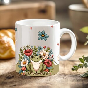 May include: White ceramic mug with a green frog sitting in a bed of pink, red, and white flowers. The mug has a white handle and a green and brown background.