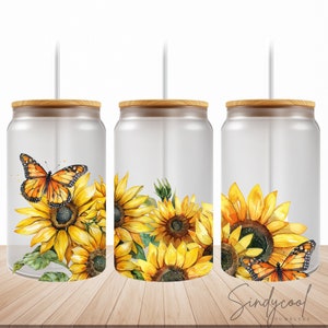 Puede incluir: Tres vasos de vidrio transparentes con tapas de bambú y pajitas. Cada vaso presenta un diseño de acuarela de girasoles y una mariposa. Los vasos están sobre una superficie de madera.