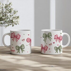 Peut inclure: Deux tasses en c&eacute;ramique blanche avec un motif de n&oelig;uds roses et verts, d'&eacute;toiles et de c&oelig;urs. Les tasses sont pos&eacute;es sur une table en bois.
