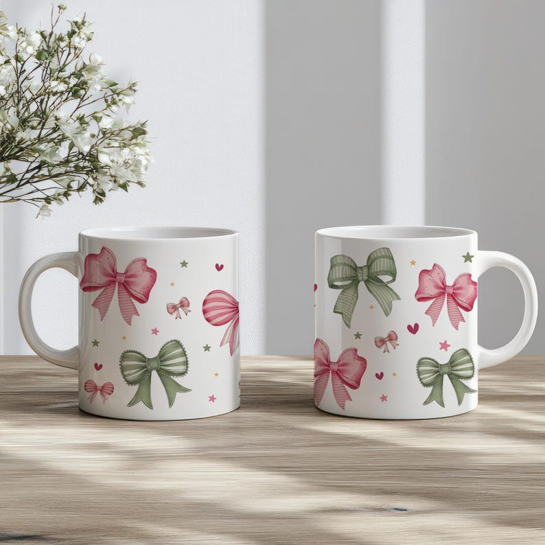 Peut inclure: Deux tasses en c&eacute;ramique blanche avec un motif de n&oelig;uds roses et verts, d'&eacute;toiles et de c&oelig;urs. Les tasses sont pos&eacute;es sur une table en bois.