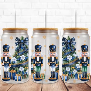 Könnte beinhalten: Drei durchsichtige Glasbecher mit einem Holzdeckel und einem Strohhalm. Jeder Becher zeigt eine Aquarell-Illustration eines Weihnachtsbaums mit einem blau-weiß gepunkteten Band, einem Nussknacker und Geschenken. Der Hintergrund ist ein weiß-goldenes Schneeflockenmuster.
