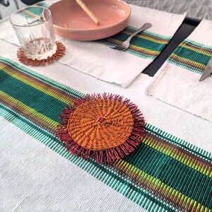 Op de afbeelding: Twee geweven placemats met groene en oranje strepen, een geweven onderzetter met franjes en een glas op een tafel.