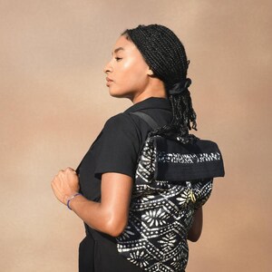 May include: A black and white patterned backpack with a flap closure. The backpack features a floral design on the back and a solid black top. The straps are black. The person is wearing a black shirt and dress.