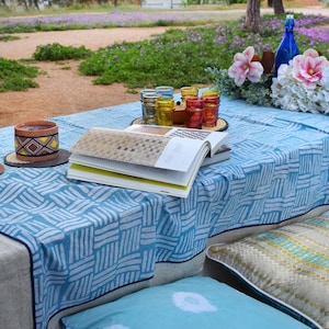 Peut inclure: Une nappe à motifs bleus et blancs avec un livre, un petit pot en céramique et quelques bouteilles en verre colorées sur le dessus. La nappe est posée sur une table avec une nappe en lin blanche en dessous. Il y a aussi quelques coussins à motifs bleus et blancs sur la table.
