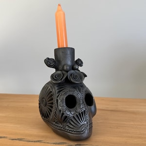 May include: A matte black ceramic skull candle holder featuring intricate carved floral and geometric patterns across its surface. The top of the skull includes a cylindrical socket holding a single, slender, solid orange taper candle.