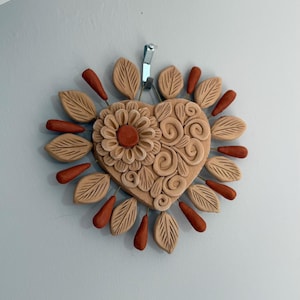 May include: A heart-shaped, tan-colored wall hanging with a floral design and leaf accents. The heart features a central flower and swirling patterns. Surrounding the heart are tan leaves and teardrop-shaped accents in a reddish-brown hue. The letter "E" is visible at the bottom.