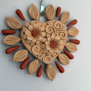 May include: A heart-shaped, handcrafted clay wall hanging. The heart features two detailed flowers and swirling designs. Surrounding the heart are clay leaves and teardrop shapes in shades of tan and rust. The letter "N" is visible at the bottom.