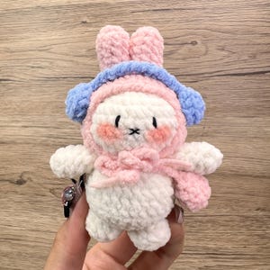 May include: A handmade plush bunny toy in white, pink, and blue. The bunny has pink ears, cheeks, and a scarf, with blue earmuffs and a hood. The toy is soft and fluffy, with black stitched eyes and a mouth.