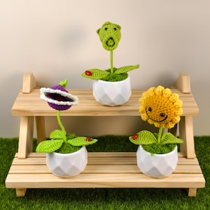 Peut inclure: Trois figurines de plantes crochetées dans des pots géométriques blancs sont posées sur une étagère en bois. Les plantes comprennent une figurine verte à bouche ouverte, une fleur violette avec une bordure blanche et un tournesol jaune souriant.