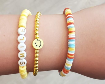May include: Three bracelets on a wrist. The first bracelet is made of yellow beads with the word "SMILE" spelled out in white letters. The second bracelet is made of gold beads with a yellow smiley face charm. The third bracelet is made of colorful beads in red, orange, yellow, blue, and white.