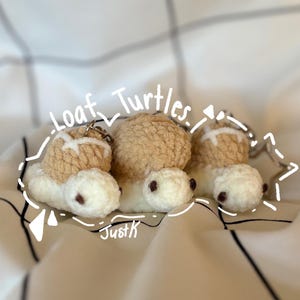May include: Three crocheted turtle keychains with white heads and tan shells resembling bread loaves. Each turtle has small dark eyes and a metal keyring attached. The text "Loaf Turtles" and "JustK" is written in white over the items.