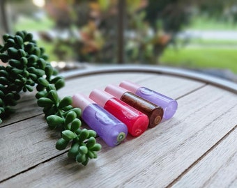 May include: Four small bottles of lip gloss with pink caps. The bottles are filled with different colored glosses: purple, pink, brown, and clear. The bottles are arranged in a row on a wooden surface.