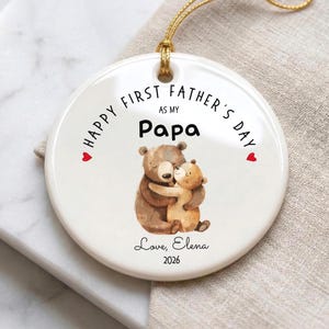 May include: A white ceramic ornament with a gold string for hanging. The ornament features the text "HAPPY FIRST FATHER'S DAY AS MY Papa" and a watercolor illustration of two hugging bears. The ornament is signed "Love, Elena 2026".