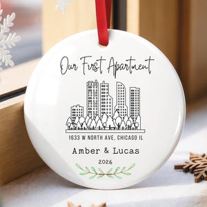 May include: A round white ceramic ornament with a red ribbon featuring a black line art city skyline, the text Our First Apartment, 1633 W NORTH AVE, CHICAGO IL, Amber & Lucas, 2026, and a small green leaf illustration at the bottom.