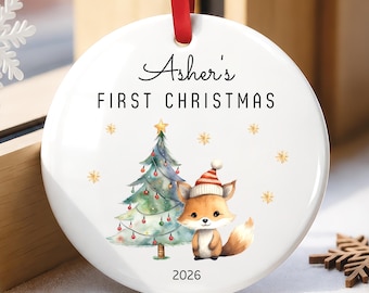 May include: A round white ceramic ornament hanging from a red ribbon. The text reads Asher's FIRST CHRISTMAS and 2026. The design features a watercolour-style Christmas tree decorated with small ornaments and a fox wearing a red and white hat.