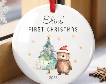 May include: A round white ceramic ornament hanging from a red ribbon. The text reads Elias' FIRST CHRISTMAS and 2026. The design features a watercolor illustration of a decorated Christmas tree with a star and a brown bear wearing a red Santa hat.