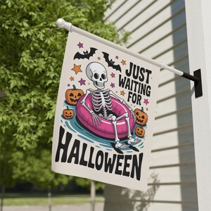 May include: A Halloween-themed flag featuring a skeleton relaxing in a pink inflatable ring. The flag includes pumpkins, bats, and stars, with the text "JUST WAITING FOR HALLOWEEN" in black. The flag is hanging outside.