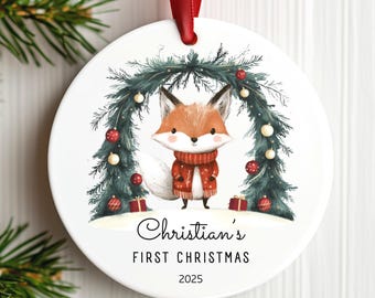 May include: A round white ceramic Christmas ornament with a red ribbon hanger. The design features a fox wearing a red sweater standing under a pine arch with red and white ornaments and gifts. Text reads: Christian's FIRST CHRISTMAS 2025.