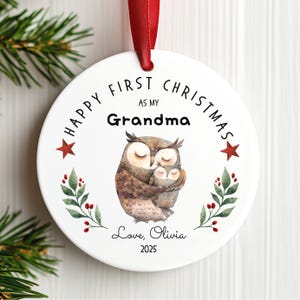 May include: A white ceramic Christmas ornament with a red ribbon. The ornament features an illustration of two owls, with text that reads "HAPPY FIRST CHRISTMAS AS MY Grandma". Additional text includes "Love, Olivia 2025".