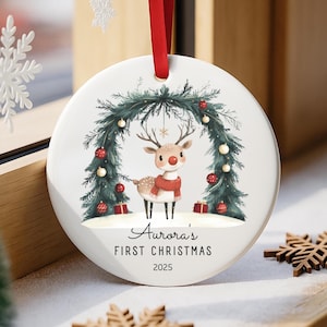May include: A white ceramic Christmas ornament with a red ribbon. The ornament features a reindeer wearing a red scarf, standing under a green arch decorated with red baubles. Text reads "Aurora's FIRST CHRISTMAS 2025".