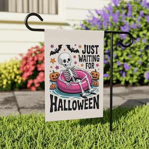 May include: A Halloween garden flag featuring a skeleton relaxing in a pink inner tube. The flag has the text "JUST WAITING FOR HALLOWEEN" with bats, stars, and pumpkins. The flag is displayed on a black metal stand in a grassy yard.