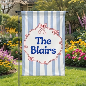May include: A personalized garden flag with blue and white vertical stripes. The flag features a white oval with the words "The Blairs" in blue script, framed by a red decorative border with bows. The flag is displayed outdoors.