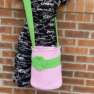 May include: A handmade pink crochet bag with a green strap and a green band around the middle. A green flower is attached to the front of the bag. The bag is displayed against a brick wall.