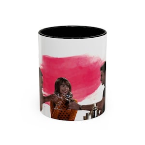 Puede incluir: Taza de cerámica blanca con interior negro. La taza presenta un gráfico de tres personas brindando con copas de vino frente a una gran forma de acuarela rosa. Las personas visten ropa informal.
