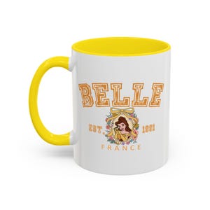 Puede incluir: Taza de cerámica blanca con interior y asa amarillos. La taza presenta la palabra "BELLE" en letras naranja, junto con una ilustración de Bella y el texto "EST. 1991 FRANCE".