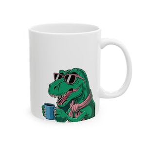Puede incluir: Taza de cerámica blanca con un T-Rex verde de dibujos animados con gafas de sol y una bufanda rosa, sosteniendo una taza azul. El dinosaurio tiene una expresión amigable. Un artículo divertido.