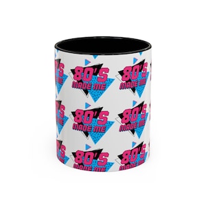 May include: A white ceramic mug with a black interior featuring a repeating pattern of pink text that reads 80'S MADE ME, layered over blue and black geometric triangle shapes with a speckled texture, creating a vibrant and colorful graphic design.