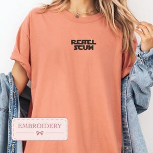 May include: A coral-colored short-sleeve t-shirt featuring the black text REBEL SCUM embroidered on the center chest. The shirt has a crew neckline and is worn with a light blue denim jacket. A rectangular label with the word EMBROIDERY is visible.