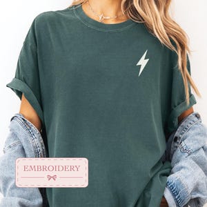 Puede incluir: Una camiseta de manga corta de color verde oscuro con un pequeño gráfico de un rayo blanco en la parte superior izquierda del pecho. La camiseta tiene cuello redondo y se lleva bajo una chaqueta vaquera azul claro. Una etiqueta dice EMBROIDERY.