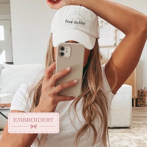 May include: A white baseball cap featuring the embroidered text free dobby in a black lowercase font on the front panel. The cap has a structured crown, a curved brim, and an adjustable strap at the back, providing a simple and clean aesthetic.