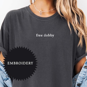 May include: A charcoal gray t-shirt with the text "free dobby" embroidered on the front. The shirt has a relaxed fit and is made of a soft, comfortable fabric. The image also includes a black starburst with the word "EMBROIDERY" in white text.