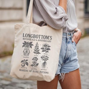 Może przedstawiać: Kremowa torba z napisem "LONGBOTTOM'S BOTANICALS & APOTHECARY" oraz ilustracjami różnych roślin. Torba zawiera również frazę "MAKERS OF RARE POTIONS". Torba jest noszona na ramieniu.