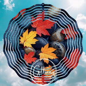 Puede incluir: Un molinillo de viento decorativo de metal con un diseño de hojas de otoño y piedras. Las hojas son rojas, naranjas y amarillas, con gotas de agua. El fondo es un cielo azul con nubes. El molinillo tiene el texto "Gillian's Design Studio".