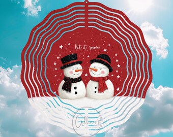 Könnte beinhalten: Ein kreisförmiger Weihnachtsschmuck mit zwei Schneemännern mit Zylindern und Schals, vor rotem Hintergrund mit dem Text „let it snow“ und weißen Sternen. Die Dekoration ist vor blauem Himmel platziert.