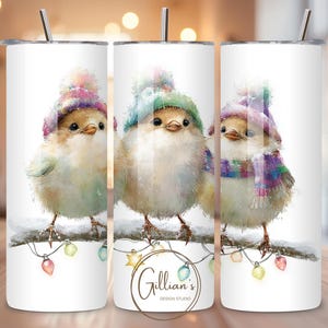 May include: White tumbler featuring three watercolor birds perched on a snowy branch, each wearing a different colored hat and scarf. The birds are surrounded by colorful Christmas lights. The tumbler has a silver straw and the text "Gillian's Design Studio".