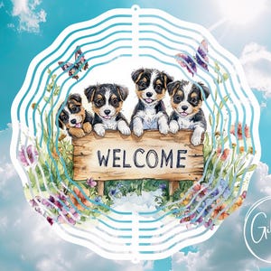 May include: A metal wind spinner with a circular design featuring four adorable puppies with brown and white fur. The puppies are holding a wooden sign that says "WELCOME". The background is a watercolor illustration of flowers and butterflies.