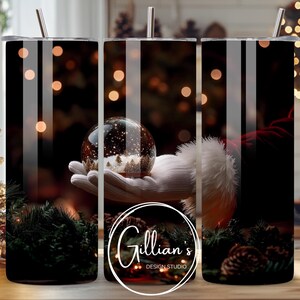 May include: Three stainless steel tumblers with a Christmas-themed design. The design features a hand holding a snow globe with a winter scene inside. The background is a blurry image of Christmas lights.