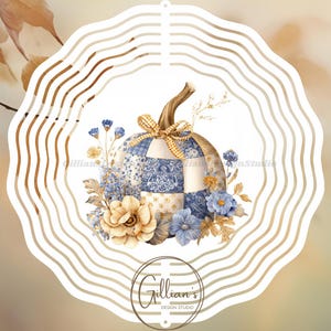 Op de afbeelding: Een aquarel illustratie van een patchwork pompoen met een gouden steel en strik. De pompoen is omgeven door blauwe en beige bloemen en bladeren, tegen een vage achtergrond. Het ontwerp is omlijst door een witte, golvende rand.