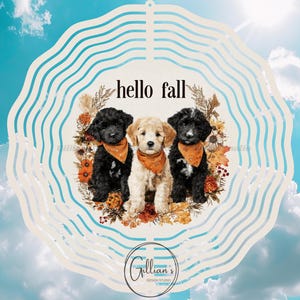 Op de afbeelding: Een decoratieve windspinner met drie schattige puppy's met oranje bandana's, omringd door herfstbladeren. De woorden "hello fall" staan boven de puppy's. De achtergrond is een blauwe lucht met wolken.