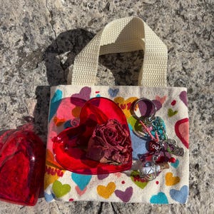 May include: A small white canvas tote bag with a red heart-shaped container with a dried rose inside. The bag has a white strap and is decorated with colorful hearts. The bag also has a keychain with a glass orb, a red heart charm, and a charm that says "LOVE".