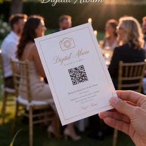 May include: A white card with gold trim and the words "Digital Album" and a camera icon. The card also has a QR code and additional text. The card is held in front of a blurred background of people at an outdoor event.